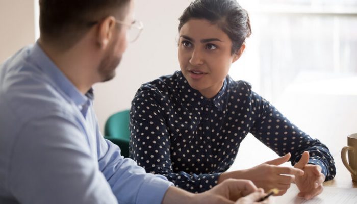Young indian female mentor coach worker talking to male coworker teaching intern having business conversation with workmate, serious hindu manager helping colleague discussing new project in office